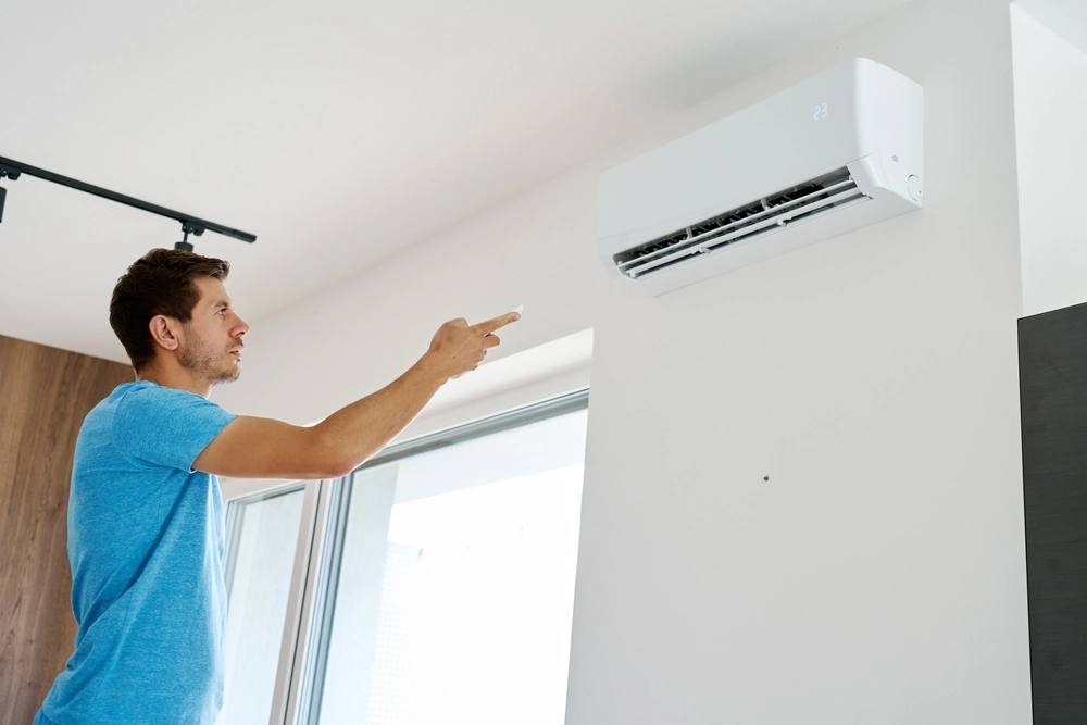 Man using remote control to adjust temperature on wall-mounted air conditioner in modern home interior. Concept of home cooling and indoor climate control during hot summer weather
