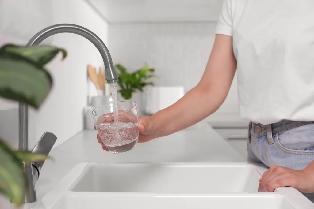 Person filling a glass with water from a kitchen faucet, countertop plants in background.