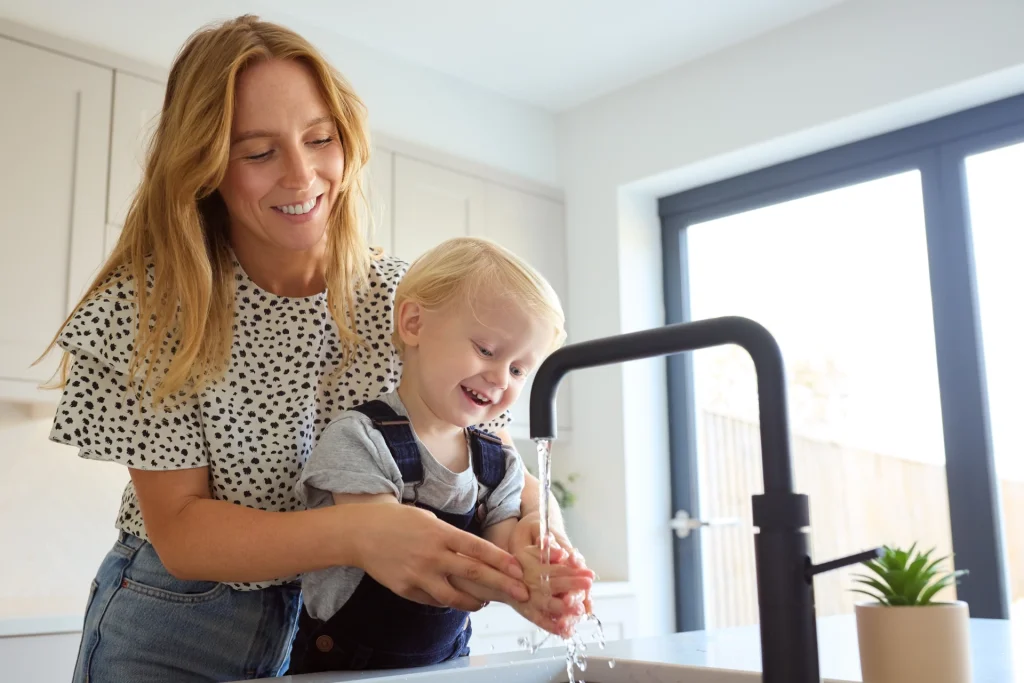 Smiling woman helps child wash hands at kitchen sink.